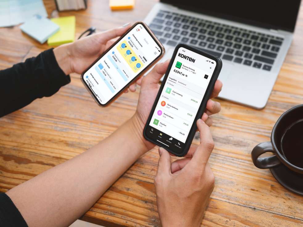 Zwei Hände halten Smartphones mit geöffneten Finanz-Apps, Laptop und Kaffee auf dem Schreibtisch.