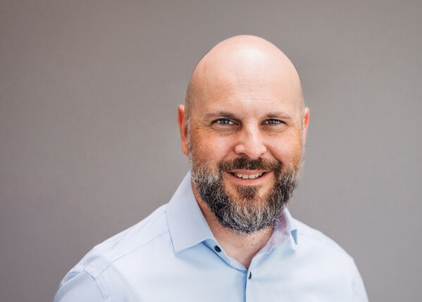 Kahlköpfiger Mann mit Bart, lächelnd, trägt ein hellblaues Hemd mit Kragen.