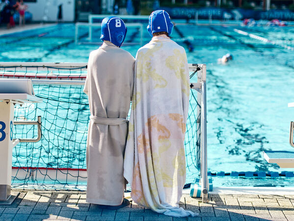 Zwei Wasserballspieler in Bademänteln und Handtüchern stehen am Rande eines Außenbeckens.