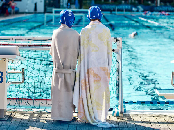 Zwei Wasserballspieler in Bademänteln und Handtüchern stehen am Rande eines Außenbeckens.