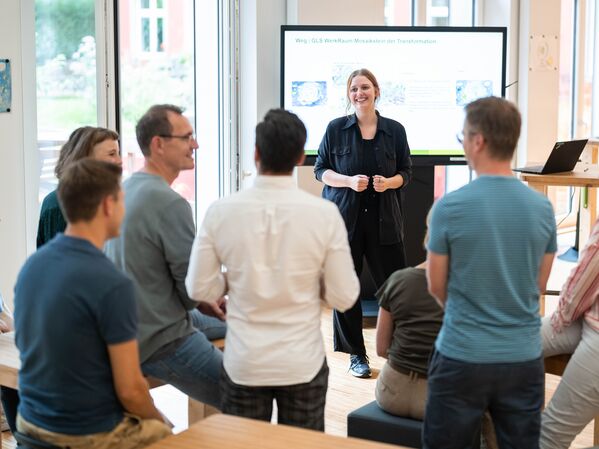 Eine Person steht vor einem Bildschirm und hält eine Präsentation. Im Vordergrund sind 7 Personen zu sehen, die zuhören und lachen.