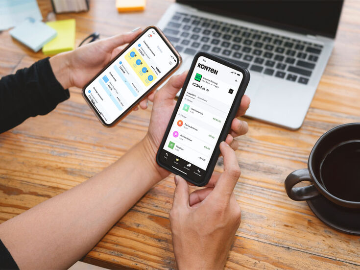 Zwei Hände halten Smartphones mit geöffneten Finanz-Apps, Laptop und Kaffee auf dem Schreibtisch.