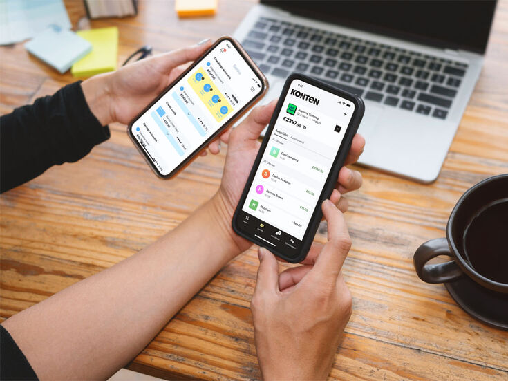 Zwei Hände halten Smartphones mit geöffneten Finanz-Apps, Laptop und Kaffee auf dem Schreibtisch.