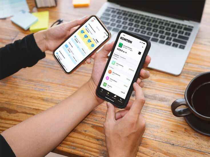 Zwei Hände halten Smartphones mit geöffneten Finanz-Apps, Laptop und Kaffee auf dem Schreibtisch.