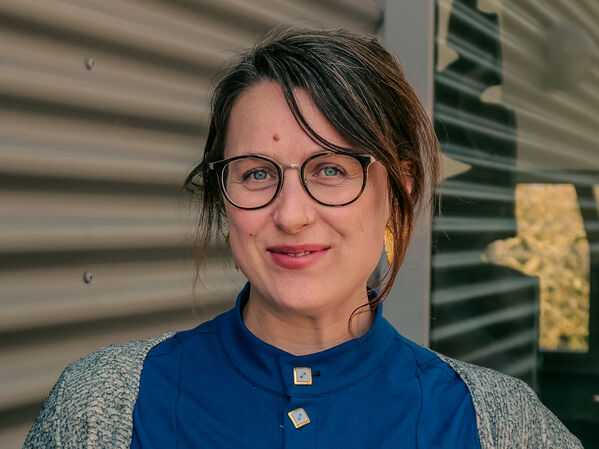 Frau mit Brille in einem blauen Kleid und grauem Pullover steht an einer Metallwand.