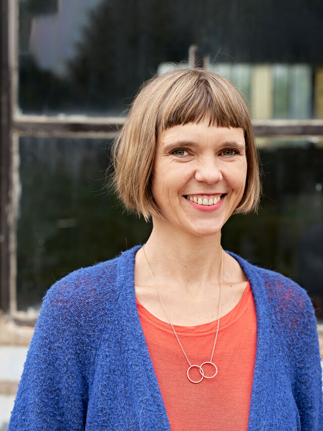 Eine lächelnde Person mit kurzen Haaren und blauer Strickjacke steht vor einem alten Fenster.