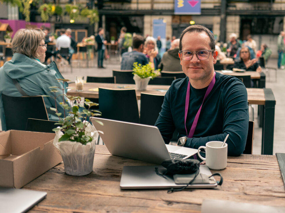 Ein Mann mit Brille sitzt an einem Tisch, auf dem ein Laptop, eine Kaffeetasse und eine Topfpflanze stehen.