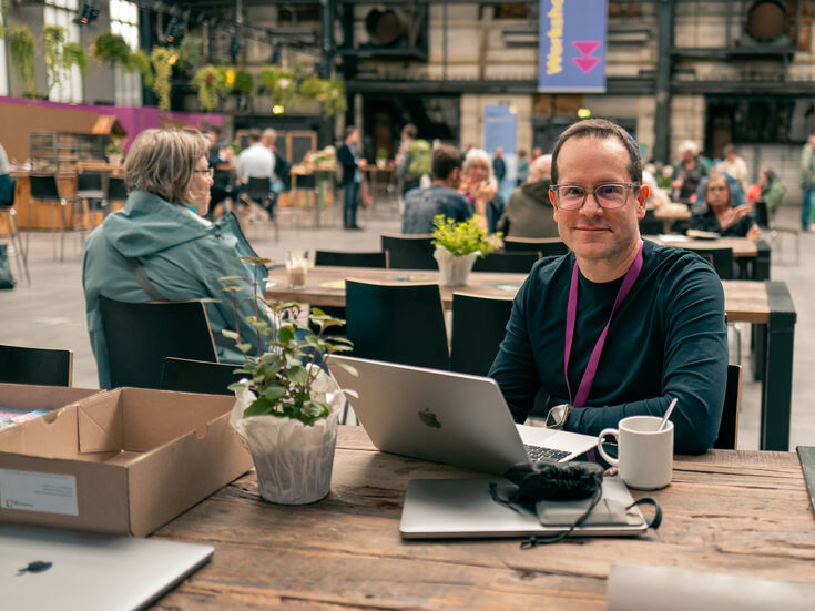 Ein Mann mit Brille sitzt an einem Tisch, auf dem ein Laptop, eine Kaffeetasse und eine Topfpflanze stehen.