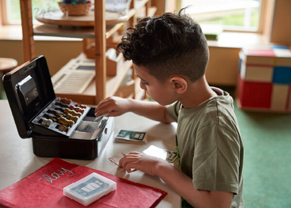 Kind sitzt an einem Tisch, vor sich eine Kasse und Spielgeld.