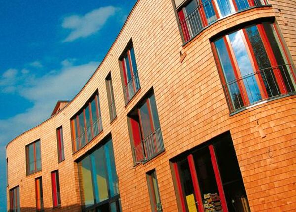 Gebogenes, modernes Gebäude mit Holzfassade und großen Fenstern unter blauem Himmel.