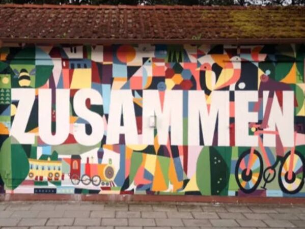 Im Hintergrund ist eine bunt bemalte Wand von einem kleinen Häuschen. Darauf steht groß in weißer Schrift "ZUSAMMEN"