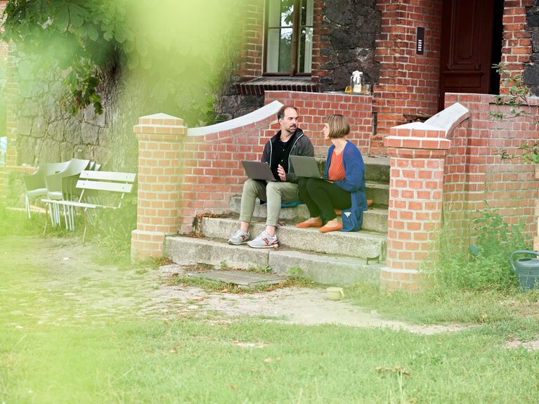 Zwei Personen sitzen auf einer gemauerten Treppe im Freien und unterhalten sich mit einem Laptop auf dem Schoß.