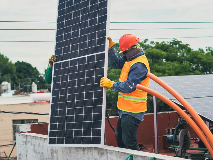 Zwei Arbeiter installieren ein Solarpanel auf einem Dach; einer trägt eine Sicherheitsweste und einen Helm.