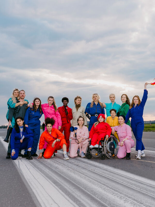 Eine bunte Gruppe von Menschen in farbenfrohen Jumpsuits posiert gemeinsam auf einem Laufsteg.