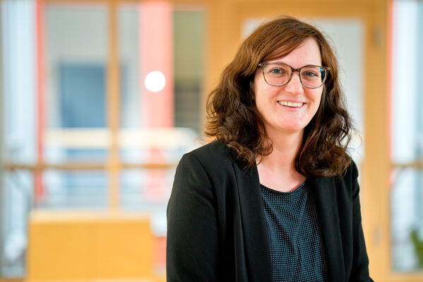 Weiblich gelesene Person mit braunem Haar und Brille, lächelnd, trägt einen schwarzen Blazer in einem Haus.