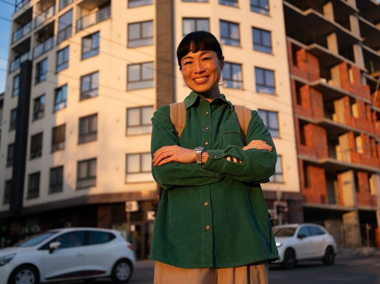 Eine Person steht lächelnd mit verschränkten Armen vor einem Wohnhaus.