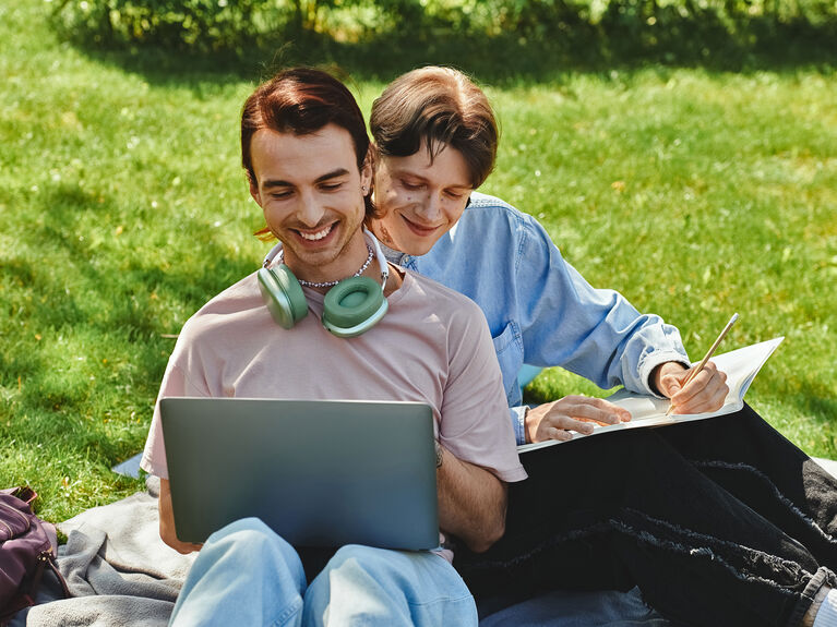 Zwei männlich gelesene Personen sitzen mit Laptop im Gras vertraut miteinander.