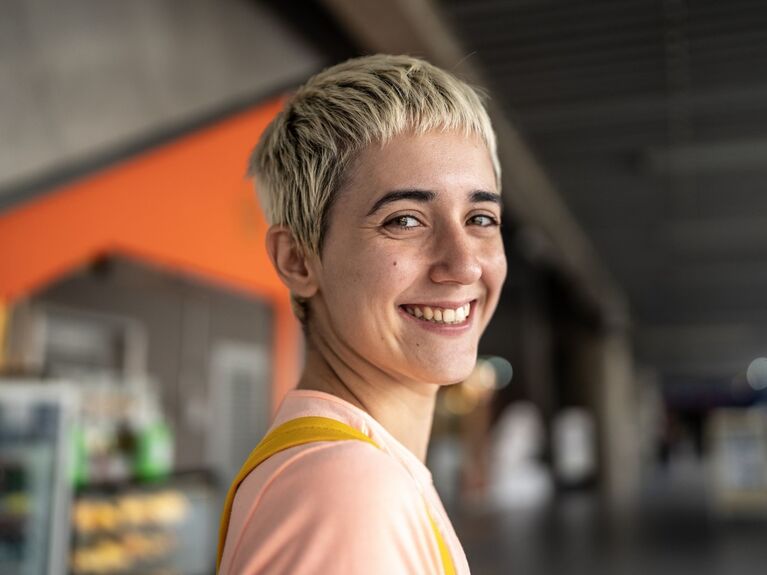 Lächelnde junge weiblich gelesene Person mit kurzen blonden Haaren.