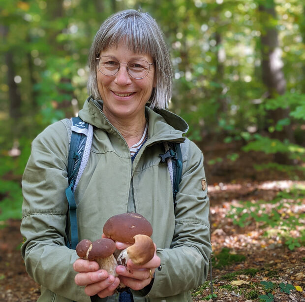 Eine Frau in einer grünen Jacke, die mehrere Pilze in der Hand hält, vor einer Waldkulisse.