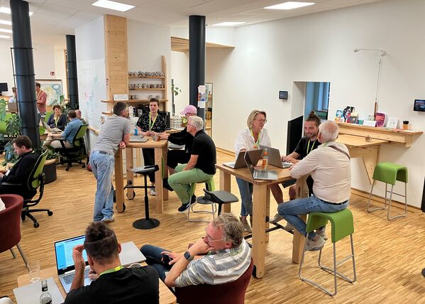 Im Bild sind 4-5 Kleingruppen zu sehen, die an niedrigen und hohen Tischen an Laptops zusammensitzen und offenbar an einer gemeinsamen Aufgabe arbeiten