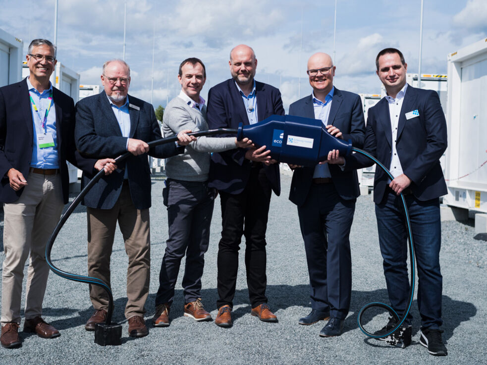 6 maennlich gelesene Personen halten ein Kabel, im Hintergrund Batteriespeicher