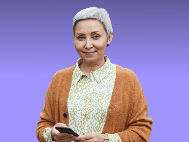 Frau mit kurzen grauen Haaren, die ein Telefon hält und ein gemustertes Hemd und eine Strickjacke trägt.